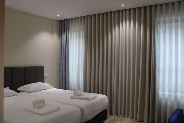 Una habitación de hotel con dos camas, cabecera oscura y un ventanal con cortinas traslúcidas.