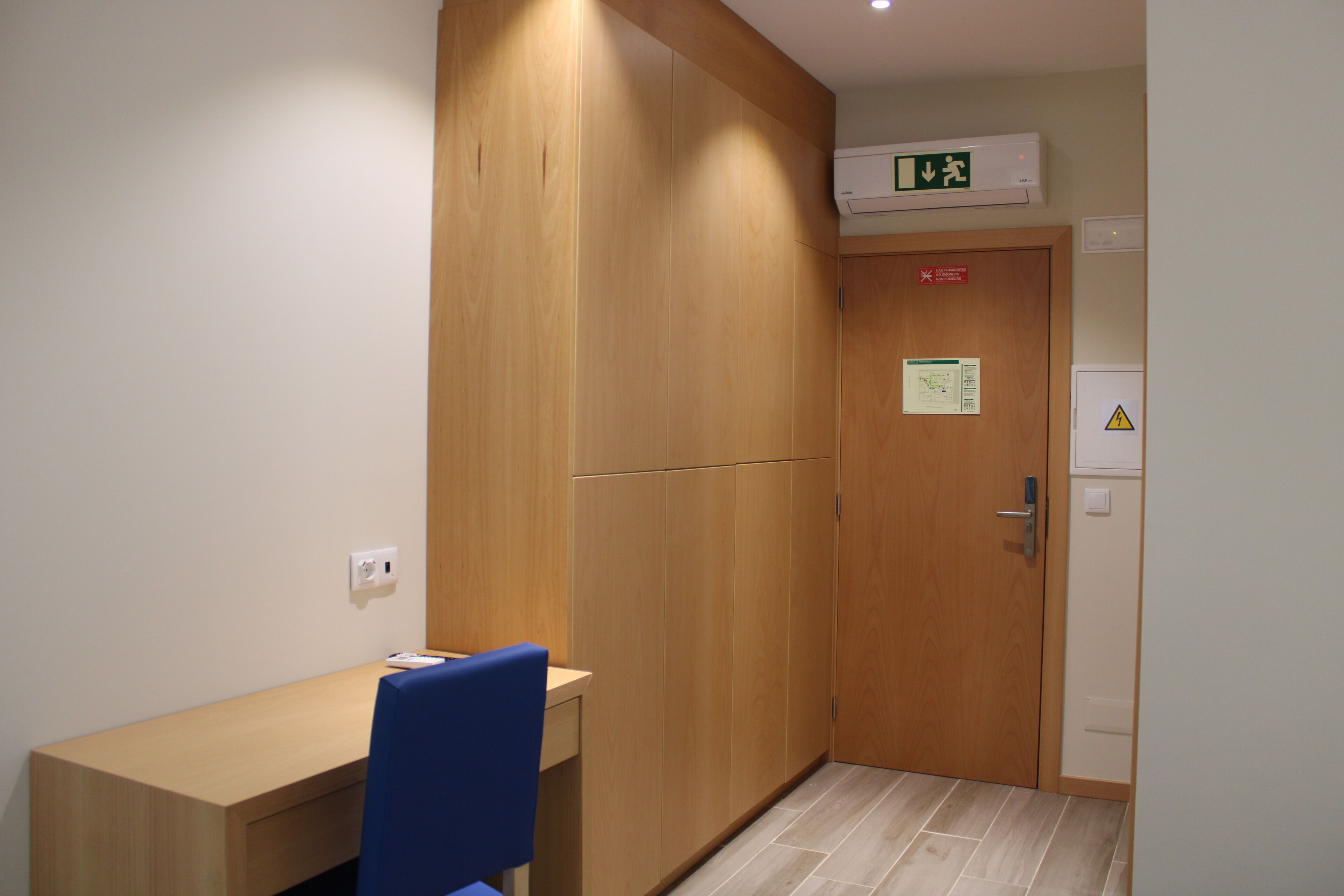 A small room with a wooden desk, a blue chair, a wooden wardrobe, and a door with a warning sign.
