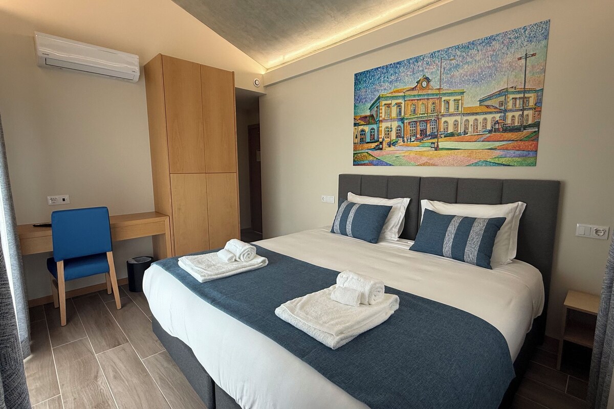 Een hotelkamer met een groot bed, twee kussens, een bureau, een stoel en een schilderij aan de muur.