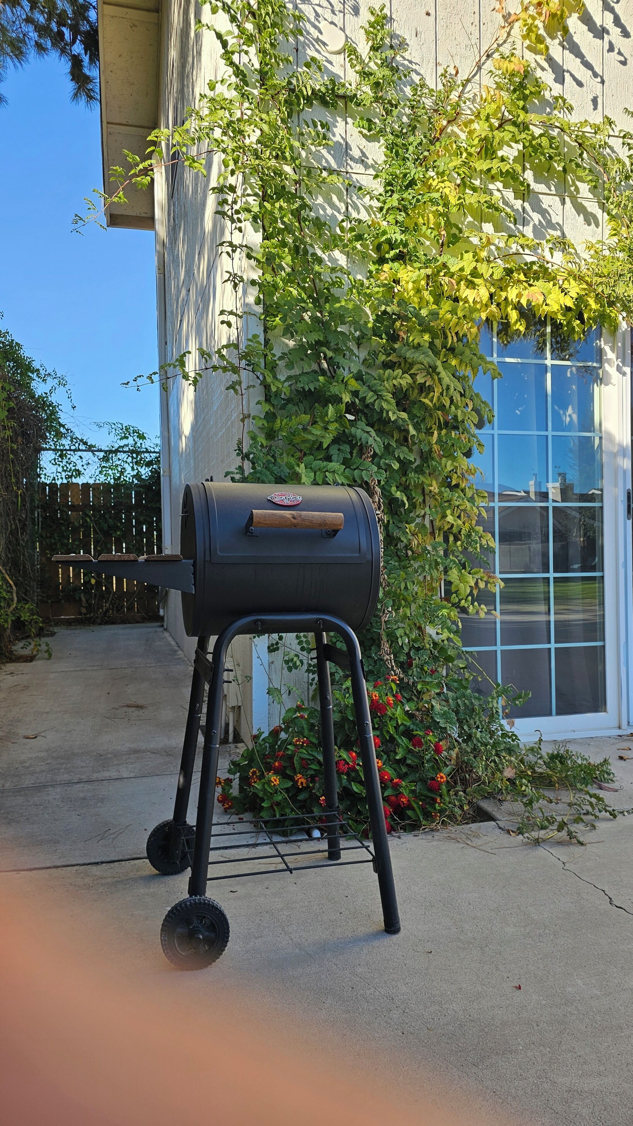A black Char-Broil grill with a metal stand, positioned on a concrete surface in front of a house with a wooden fence and a window.