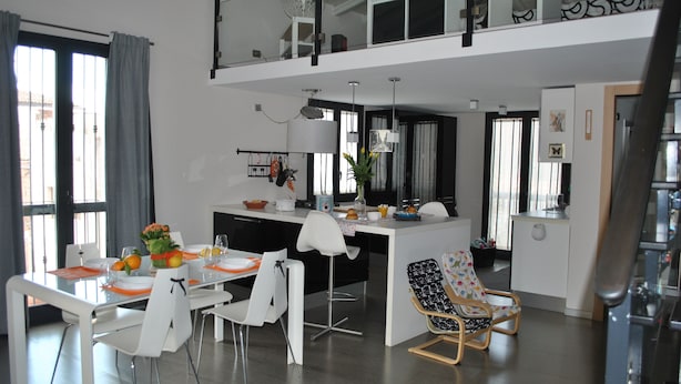 A modern kitchen with a central island, white dining area with chairs, and a view of the city through large windows. - The Odeon Terrace (Catania)