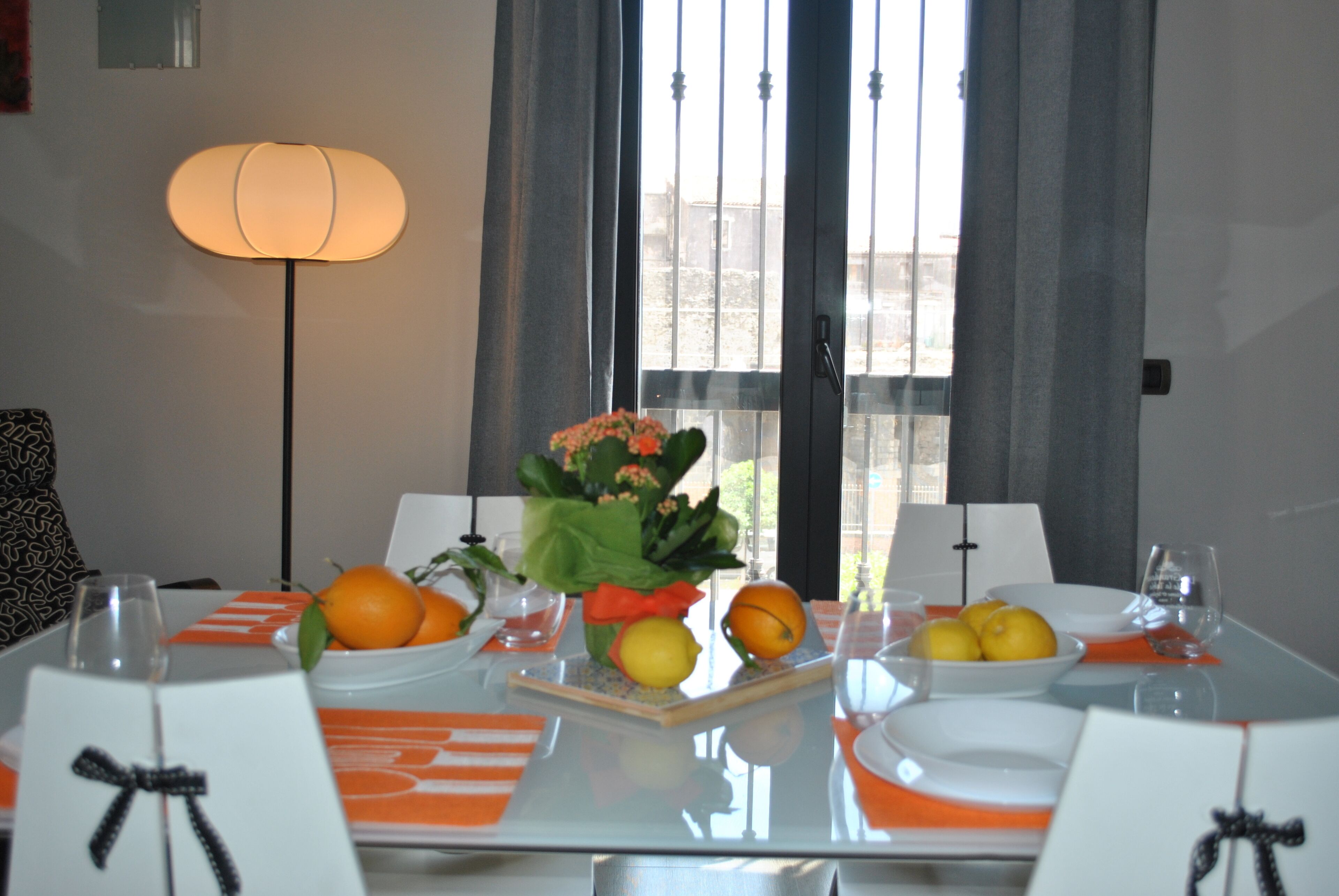 Una mesa de comedor dispuesta con frutas anaranjadas, un florero con flores y una lámpara al fondo.