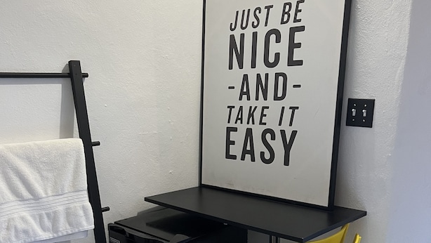Ein kompakter Raum mit schwarzem Schreibtisch, gelbem Stuhl und kleinem, schwarzem Kühlschrank. Ein Handtuchhalter mit weißen Handtüchern ist sichtbar, und an der Wand hängt ein motivierender Poster.