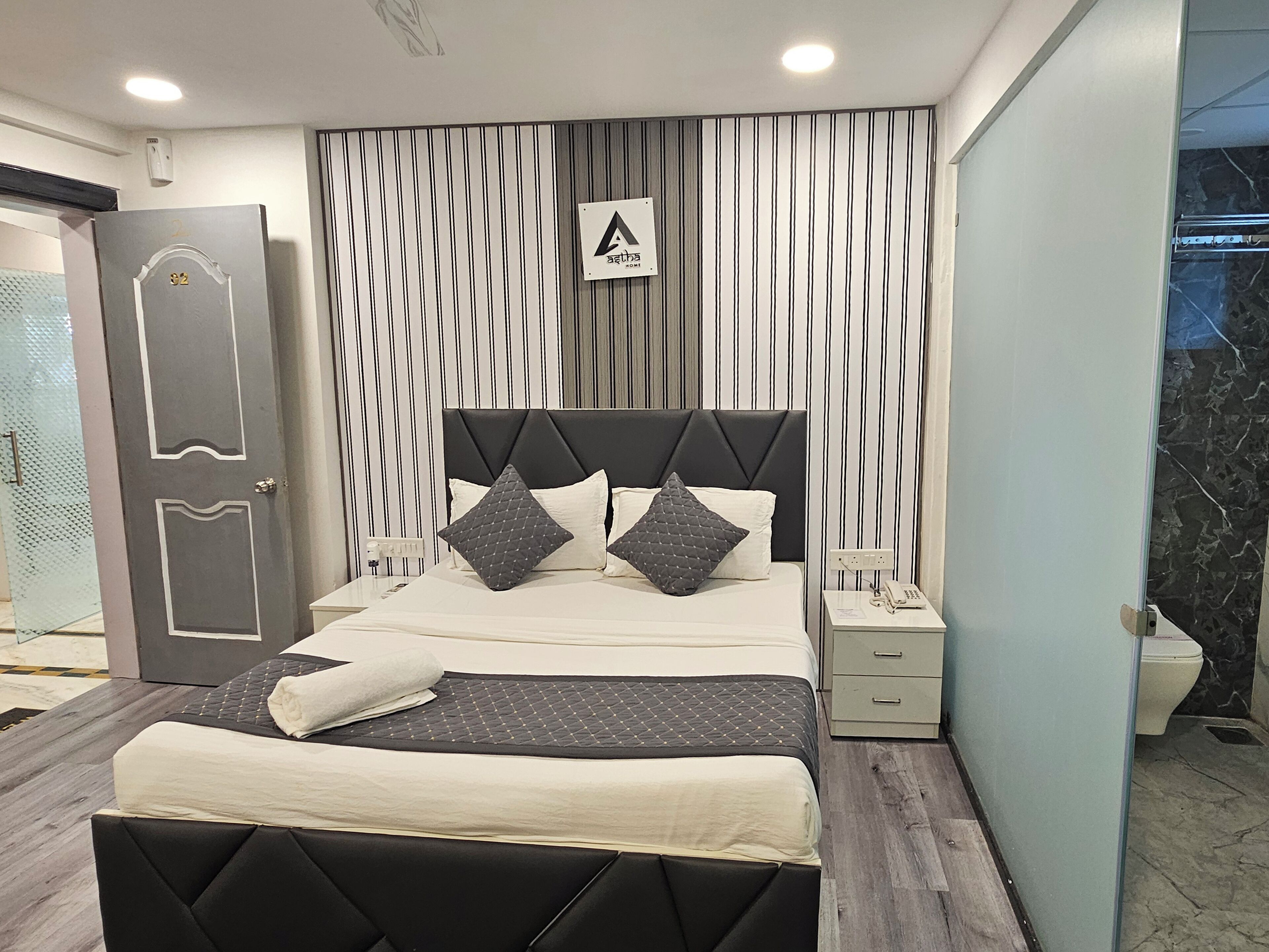 A modern bedroom with a large bed, a nightstand, a wall-mounted phone, and a door with a number plate.