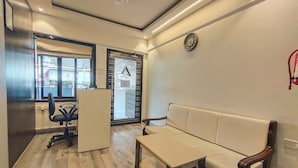 A room with a wooden floor, a white sofa, a small table, a black office chair, and a window with blinds. - Astha Kandivali Service Apt (Mumbai)