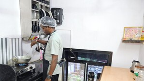 A kitchen with a stove, a person cooking, and a refrigerator. - Astha Kandivali Service Apt (Mumbai)