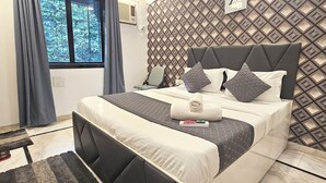 A neatly made bed with a headboard, a window with curtains, a patterned wall, and a small table with a chair. - Astha Kandivali Service Apt (Mumbai)