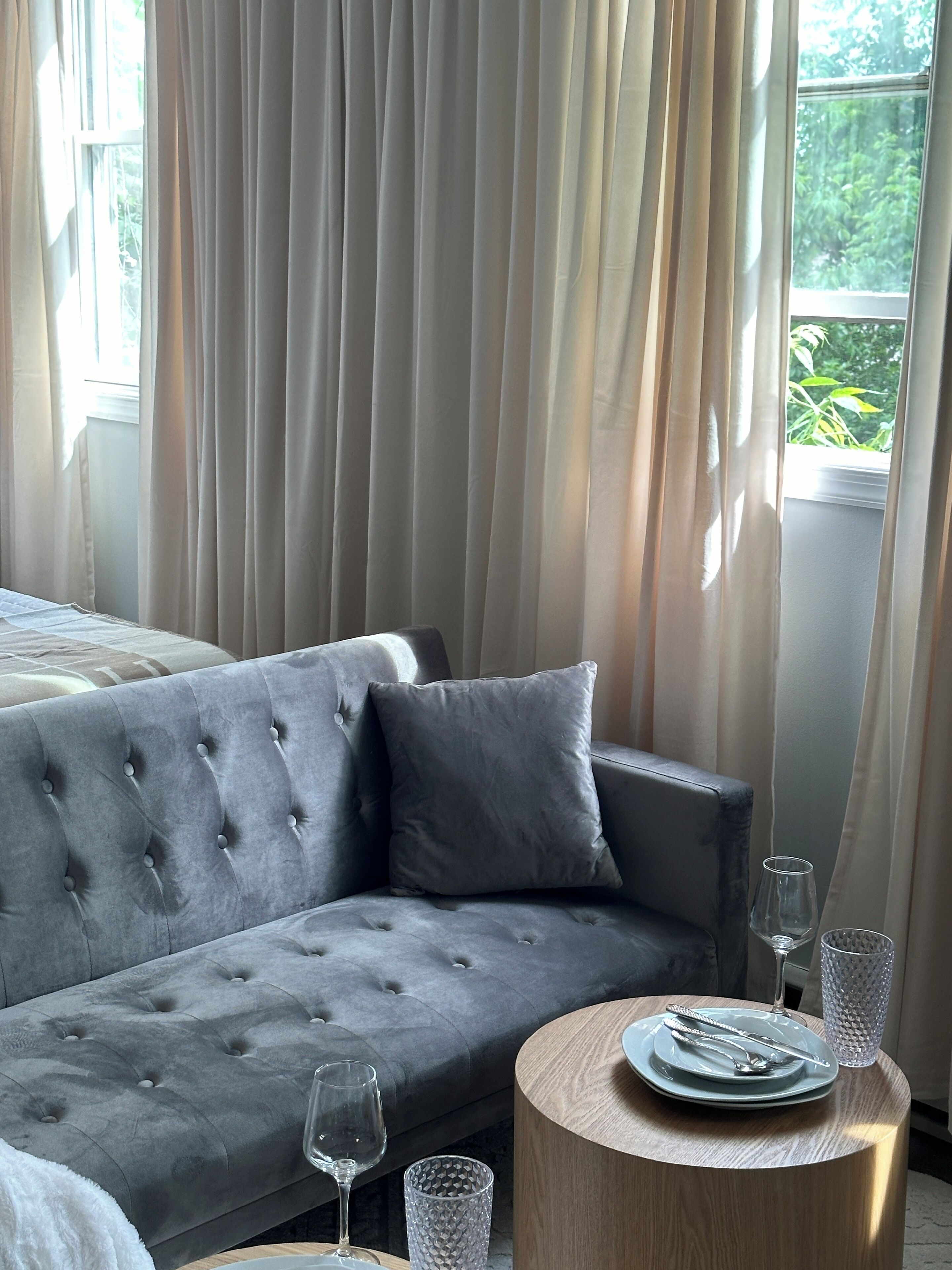 Quarto moderno com sofá cinza estofado, mesa redonda de madeira com prato e talheres, e janela com cortinas translúcidas.