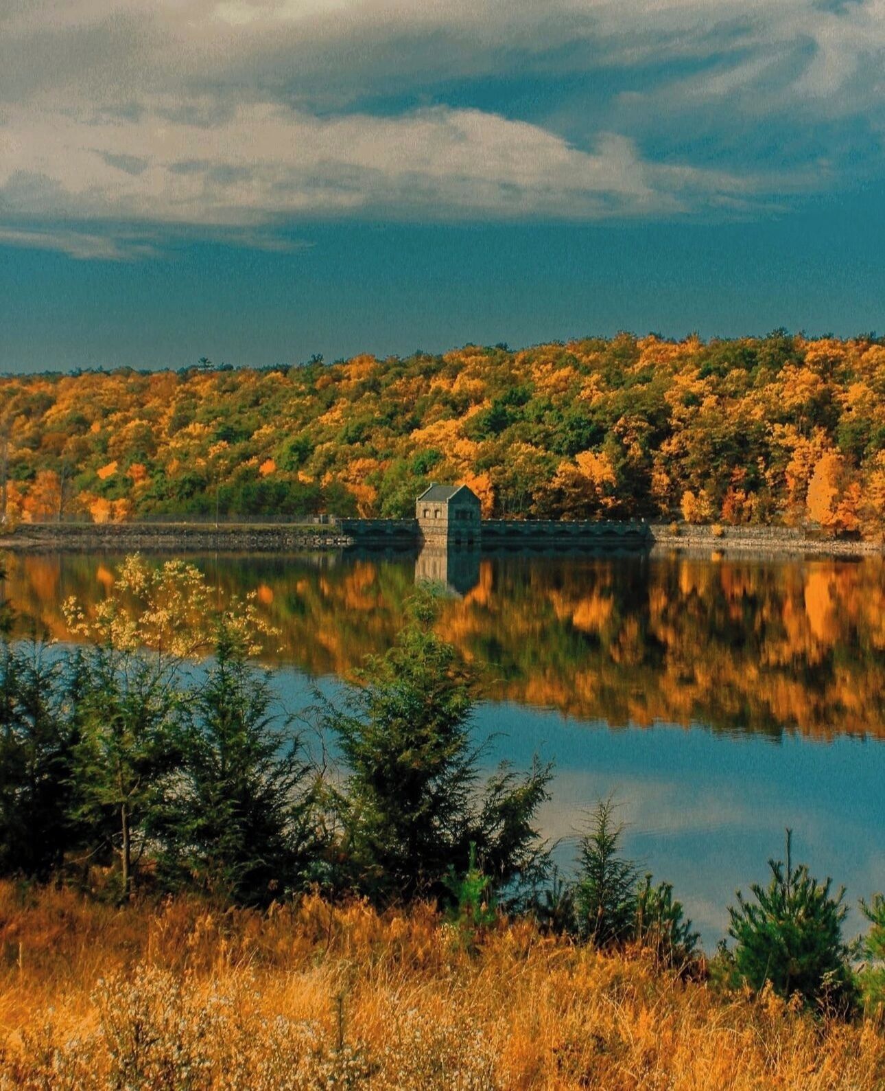 A serene landscape with a small stone structure by a calm lake, surrounded by autumn foliage.