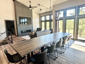 Uma sala de jantar espaçosa com uma mesa comprida e várias cadeiras, lareira e grandes janelas com vista para o exterior.