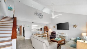 A spacious living room with a white sofa, a coffee table, and a TV. There's a staircase with wooden steps leading to another room.