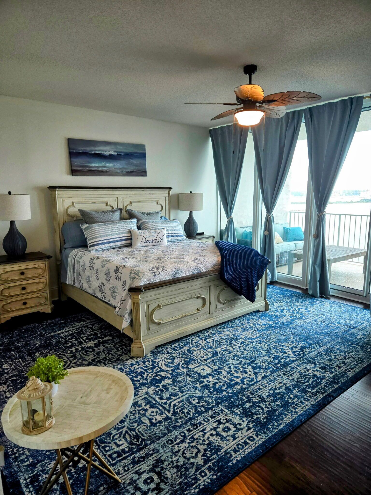 A bedroom with a bed, bedside tables, a nightstand, a round table with a lantern, and a ceiling fan.
