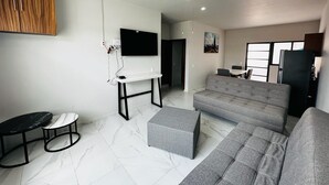 Een moderne woonkamer met een grijze bank, een klein rond tafeltje, een aan de muur bevestigde televisie en een plafondventilator.
