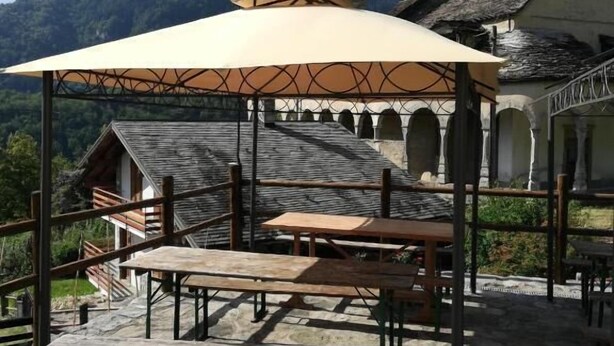 A stone patio with wooden benches and a canopy, overlooking a mountain and a traditional building with a bell tower. - Rifugio Monte Zeus (Premia)