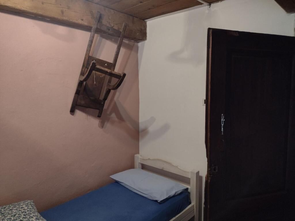 Ein kleiner Raum mit einem Einzelbett, einem Holzstuhl, einer Tür und einer an der Wand befestigten Leiter.