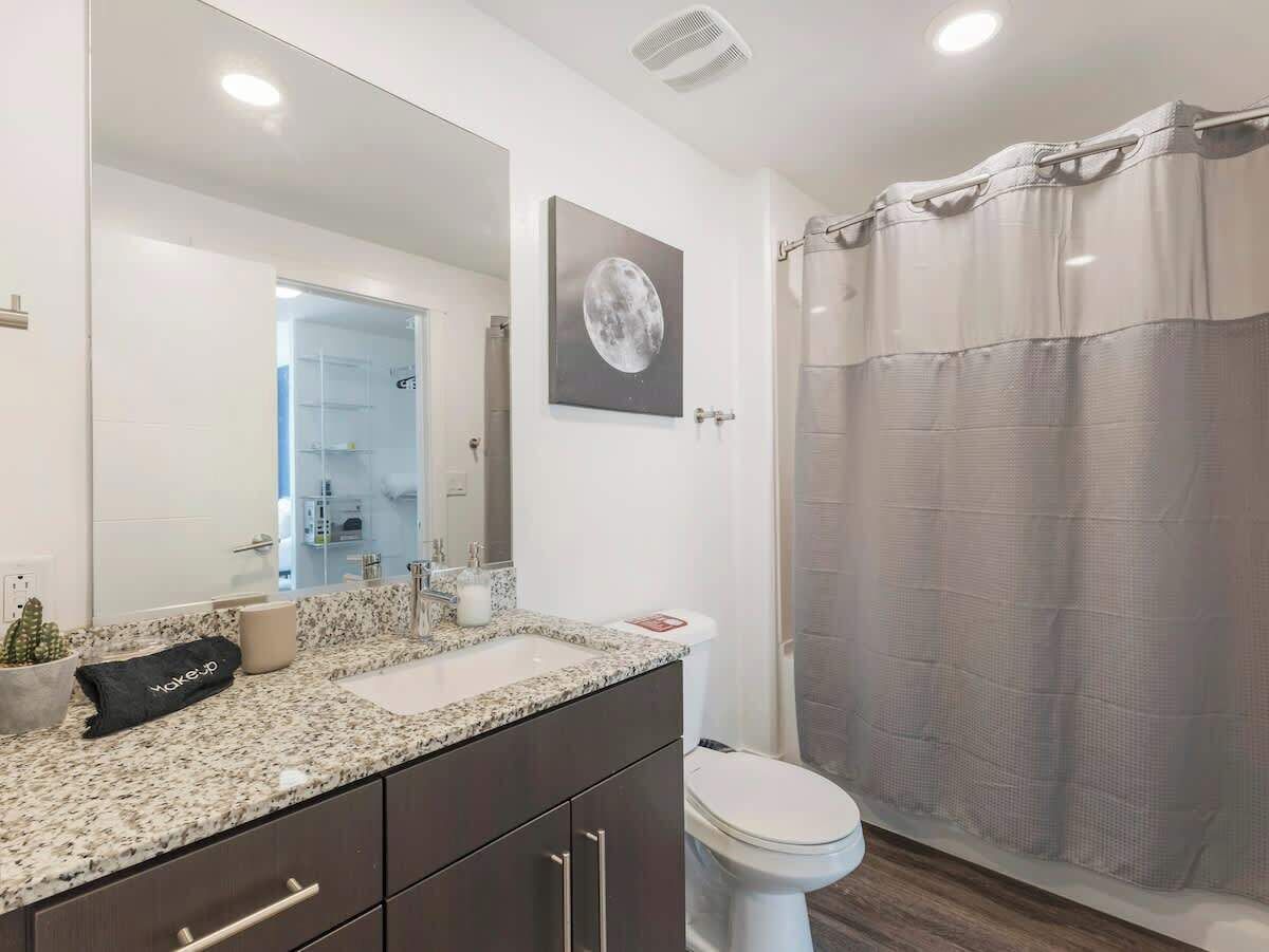 Une salle de bain moderne avec un comptoir en granit, des toilettes blanches, un rideau de douche et un grand miroir.