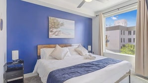 Une chambre d’hôtel avec un lit, une table de chevet, un ventilateur de plafond, une fenêtre avec des rideaux et un tableau au mur.