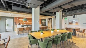 Un espacio de oficina moderno con un amplio escritorio de conferencias de madera rodeado de sillas verdes, una cocina con salpicadero de diseño estampada tipo espiga y un cuadro en la pared.