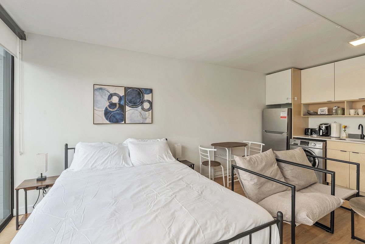 Une chambre d’hôtel moderne comprenant un lit, une petite table à manger, une chaise, un coin cuisine et une œuvre d’art accrochée au mur.