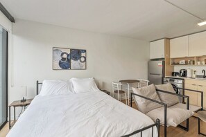 Habitación de hotel moderna con cama, una pequeña mesa de comedor, una silla, kitchenette y una obra de arte en la pared.