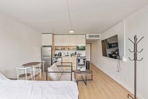 Habitación de hotel compacta con cama, pequeña mesa de comedor con sillas, kitchenette con estufa y microondas, y perchero.
