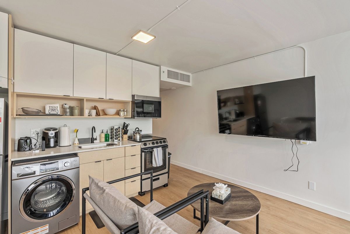 A compact kitchen with a washing machine, oven, microwave, and a flat-screen TV mounted on the wall.
