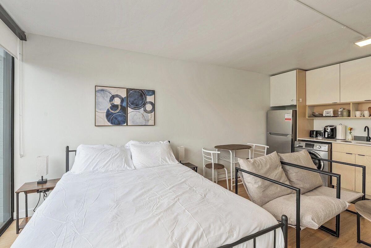 Moderni hotellihuone, jossa on sänky, pieni ruokapöytä, tuoli, minikeittiö ja seinälle kiinnitetty taideteos.