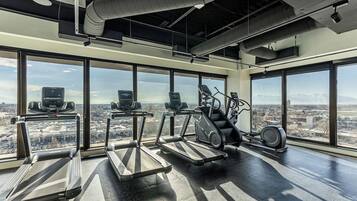 A gym with treadmills, elliptical machines, and stationary bikes, large windows with a city view.