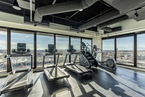 A gym with treadmills, elliptical machines, and stationary bikes, large windows with a city view.