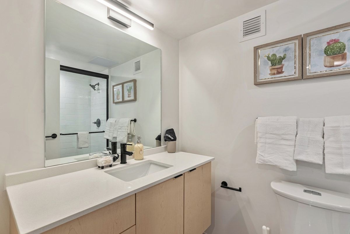 Une salle de bain moderne avec un grand miroir, un lavabo blanc et des toilettes. Des tableaux encadrés ornent le mur, et un porte-serviettes est également présent.