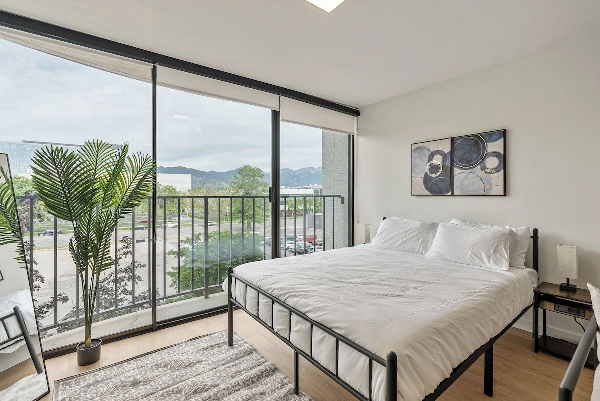 Une chambre moderne avec un grand lit, une vue sur la ville grâce à de grandes fenêtres du sol au plafond, une plante en pot et un tableau au mur.