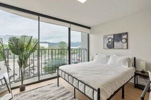 A modern bedroom with a large bed, a view of the city through floor-to-ceiling windows, a potted plant, and a painting on the wall.