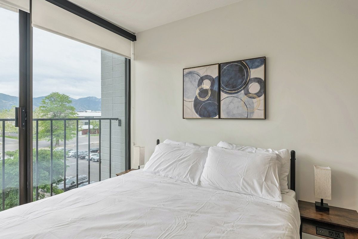 Une chambre avec une grande fenêtre, un lit recouvert de draps blancs, une table de chevet et une œuvre d’art abstraite accrochée au mur.