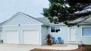 Een lichtblauwe gelijkvloerse woning met garage, een klein terras met twee blauwe Adirondack-stoelen, en een verzorgd gazon.