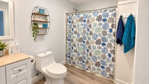 Un baño con inodoro, estante con artículos de aseo y cortina de ducha estampada con motivos de conchas marinas.
