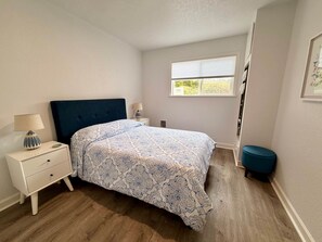 Un dormitorio con cama, mesita de noche, lámpara, ventana y un reposapiés azul.