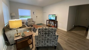 Een woonkamer met een bank, fauteuil, tafel, lamp, televisie en een gestreept voetenbankje.