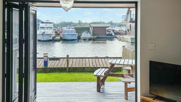 Una stanza con una porta scorrevole in vetro che dà su una terrazza con televisore e angolo salotto, affacciata su un porto turistico con barche.