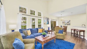 A spacious living room with a blue carpet, a blue sofa, and a wooden coffee table. There are two windows with white curtains, a dining area with a table and chairs, and a painting on the wall. - Townhome Perfectly Located Near Clubhouse (Kissimmee)