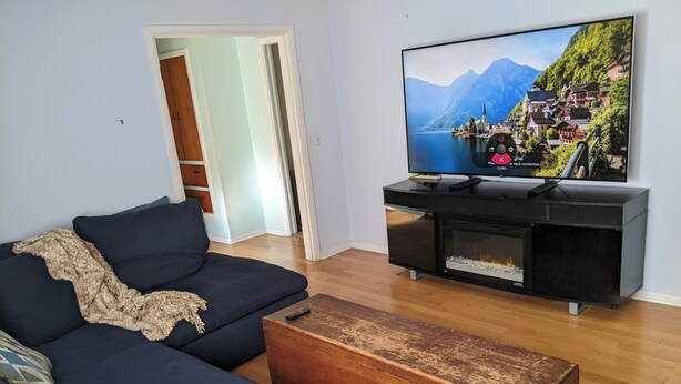 A living room with a dark blue sofa, a wooden coffee table, and a television displaying a scenic mountain village. - UM Football - 5 blocks to Stadium (Ann Arbor)