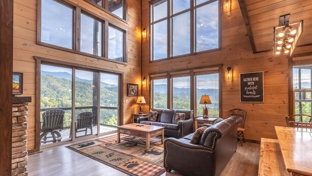 Un interior espacioso de madera con grandes ventanales que ofrecen vistas a la montaña, una zona de estar acogedora con chimenea y una mesa de comedor.