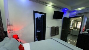A modern hotel room with a bed, a red chair, a TV, and a bathroom visible through an open door.