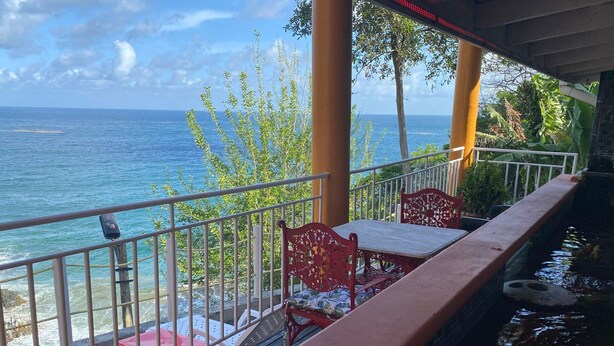A balcony with a view of the ocean, featuring a table and chairs, a small water feature, and a railing.