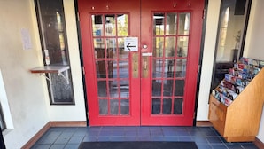 Una puerta roja con un felpudo que dice 'Welcome', una puerta de cristal y un mueble para revistas a la derecha.