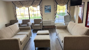 Una sala de estar con sofás color beige, una mesa de centro y dos sillones. Hay ventanas grandes con cortinas, un televisor de pantalla plana montado en la pared y una pequeña alfombra en el suelo.