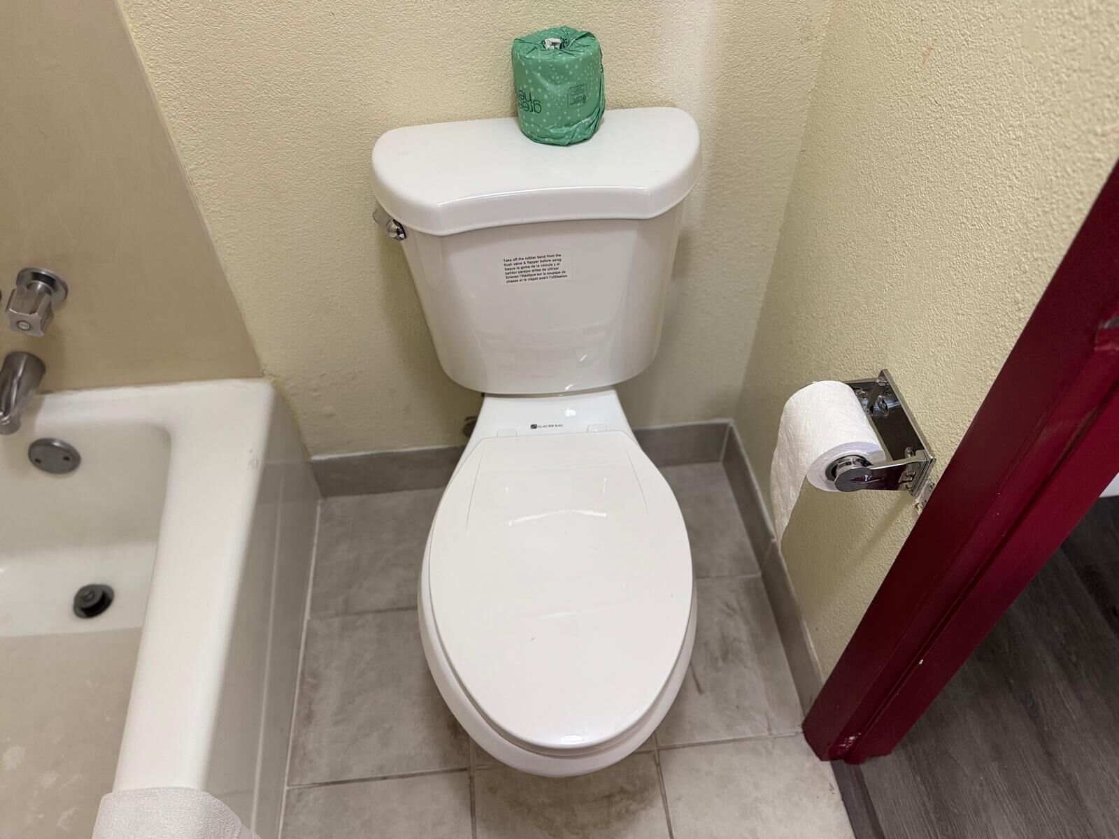 Inodoro blanco con un dispensador de papel higiénico verde, un rollo de papel higiénico blanco y una bañera blanca con un grifo.