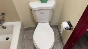 Inodoro blanco con un dispensador de papel higiénico verde, un rollo de papel higiénico blanco y una bañera blanca con un grifo.