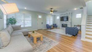 Un salon spacieux avec un grand canapé d’angle, une table basse et deux fauteuils. On y trouve une cheminée, un téléviseur fixé au mur et un ventilateur de plafond.