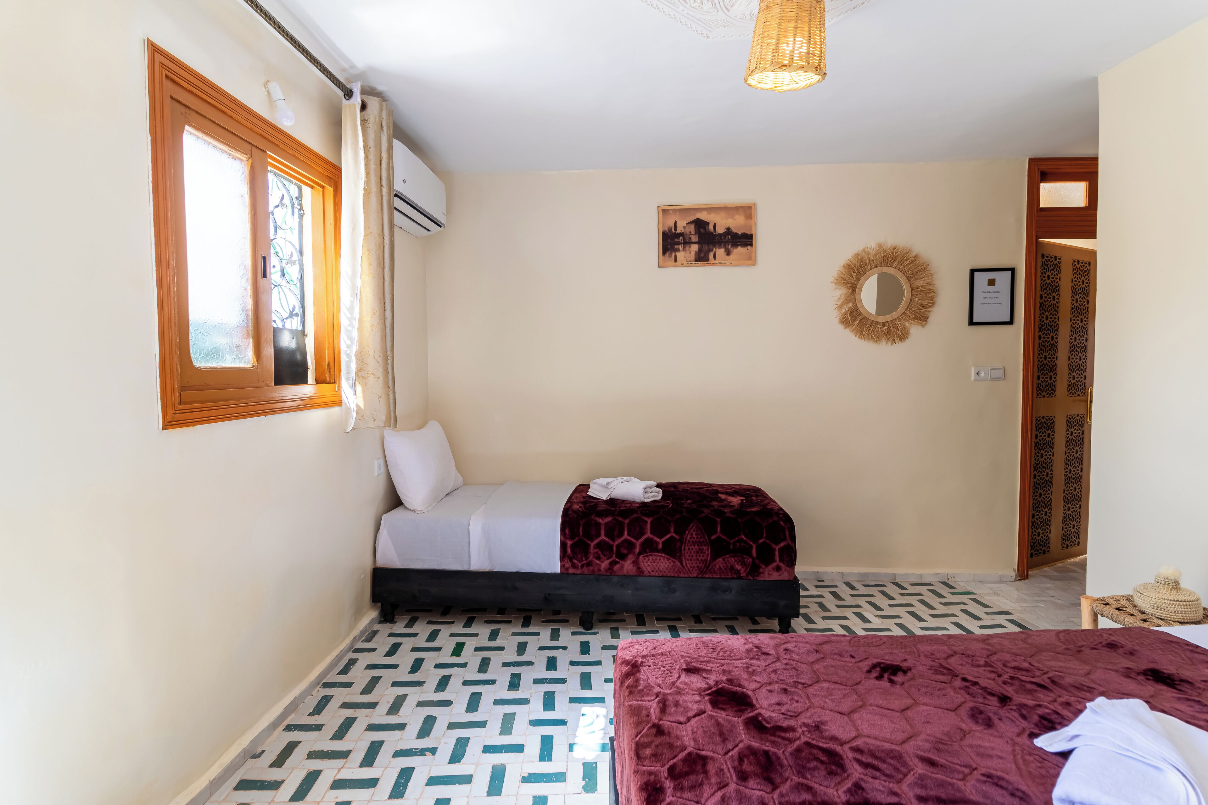 A bedroom with two beds, patterned flooring, a window with curtains, a wall-mounted air conditioner, a framed picture, and a decorative mirror.