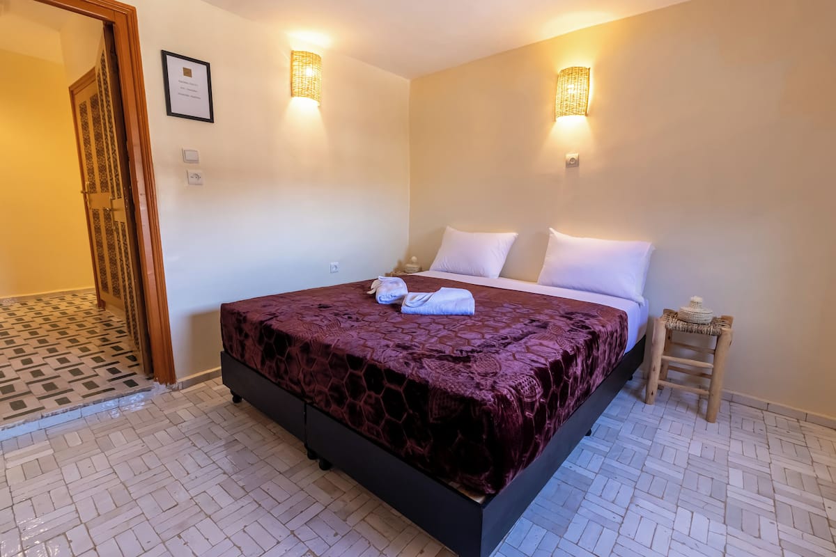 Une chambre d’hôtel avec un grand lit, deux lampes fixées au mur, une petite table de chevet et un tabouret en bois.